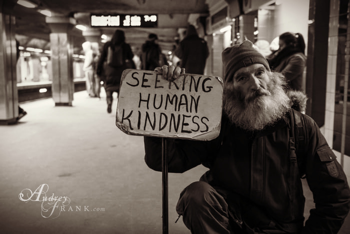 An elderly man sitting in a subway, holding a sign that says "Seeking Human Kindness"
